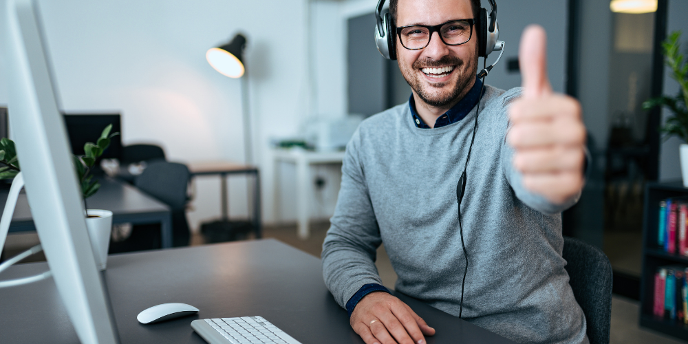 smiling man at desk wearing headphones and giving a thumbs up