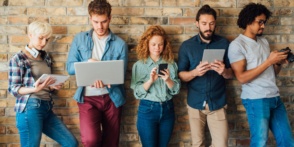 5 millennials on different devices leaning against brick wall